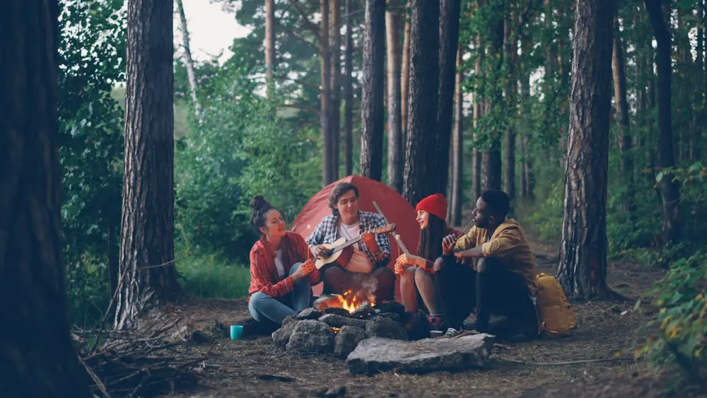 Cuatro amigos sentados alrededor de una hoguera en un bosque; uno toca la guitarra y detrás hay una tienda de campaña roja.