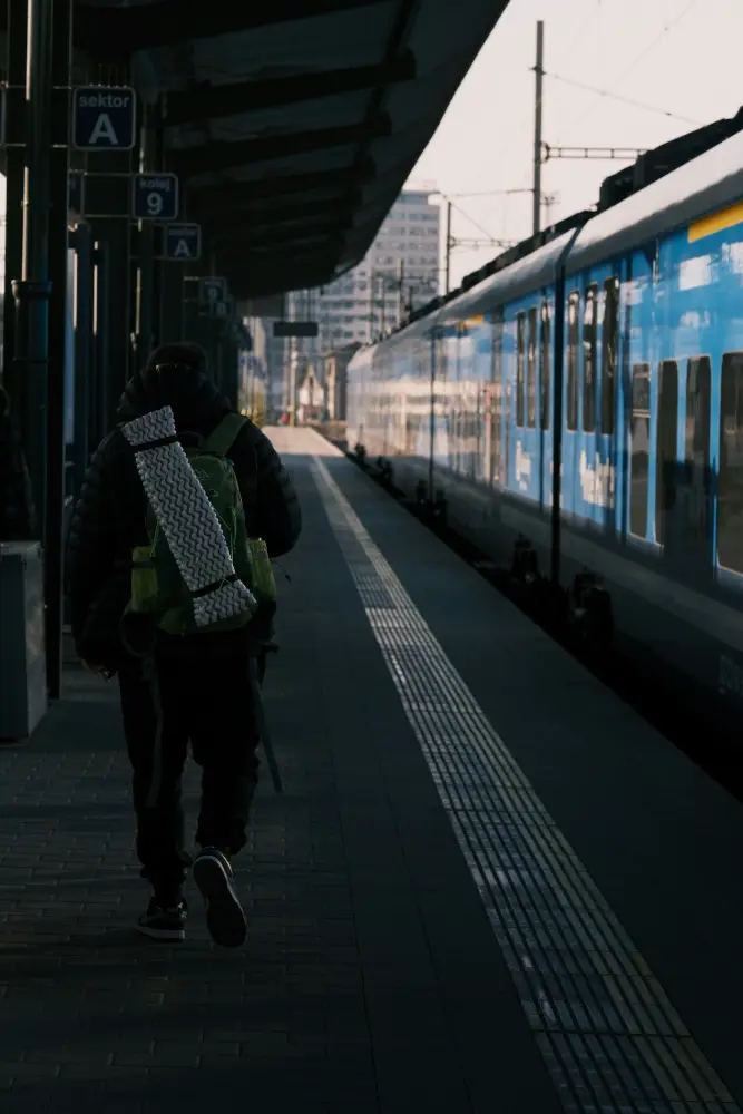 Una persona con mochila grande camina por un andén junto a un tren azul y blanco, con letreros de sectores colgados del techo.