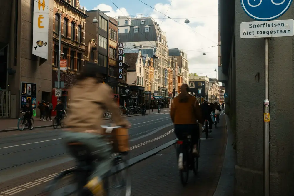 Varias personas van en bici por una calle con edificios antiguos de ladrillo; en la pared hay un cartel en neerlandés.