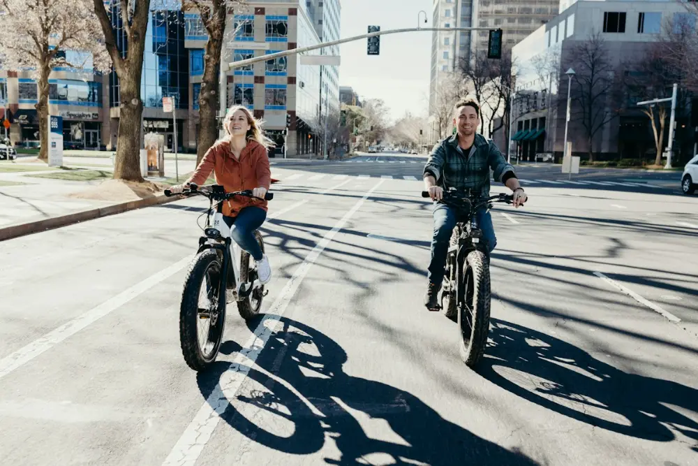 Un hombre y una mujer van en bicicleta eléctrica por una avenida soleada de una ciudad con edificios altos.