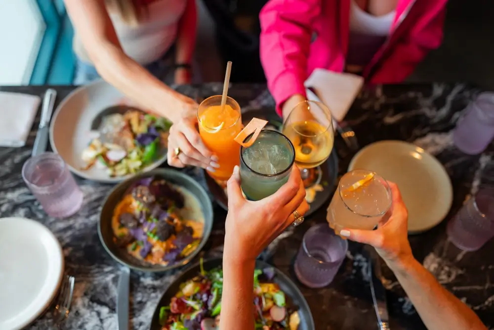 Vista desde arriba de una mesa con platos de comida; varias manos levantan vasos con bebidas para brindar.