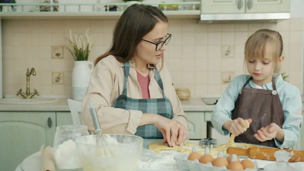 Una madre con gafas y una niña rubia, las dos con delantal, preparan masa sobre una mesa con harina y huevos.