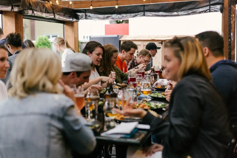 Un grupo grande de amigos come y charla alrededor de una mesa larga en una terraza cubierta, con bebidas y platos.