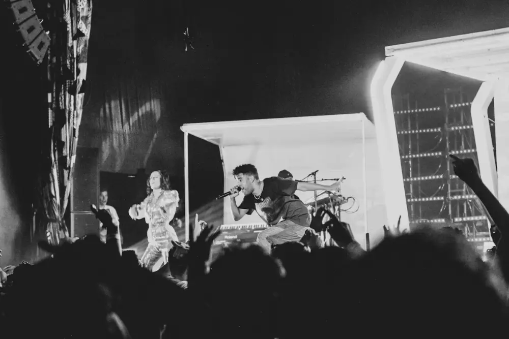 Foto en blanco y negro de un concierto: un cantante con micrófono y una mujer bailando en el escenario; el público levanta las manos.