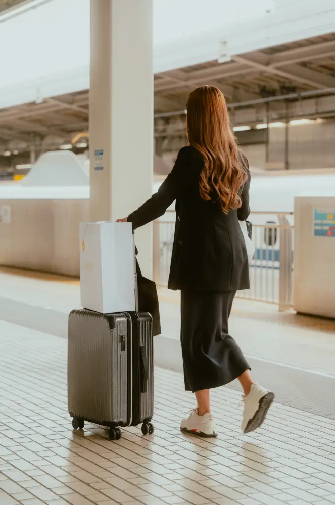 Una mujer de pelo largo y abrigo negro camina por el andén de una estación arrastrando una maleta gris con una bolsa blanca encima.