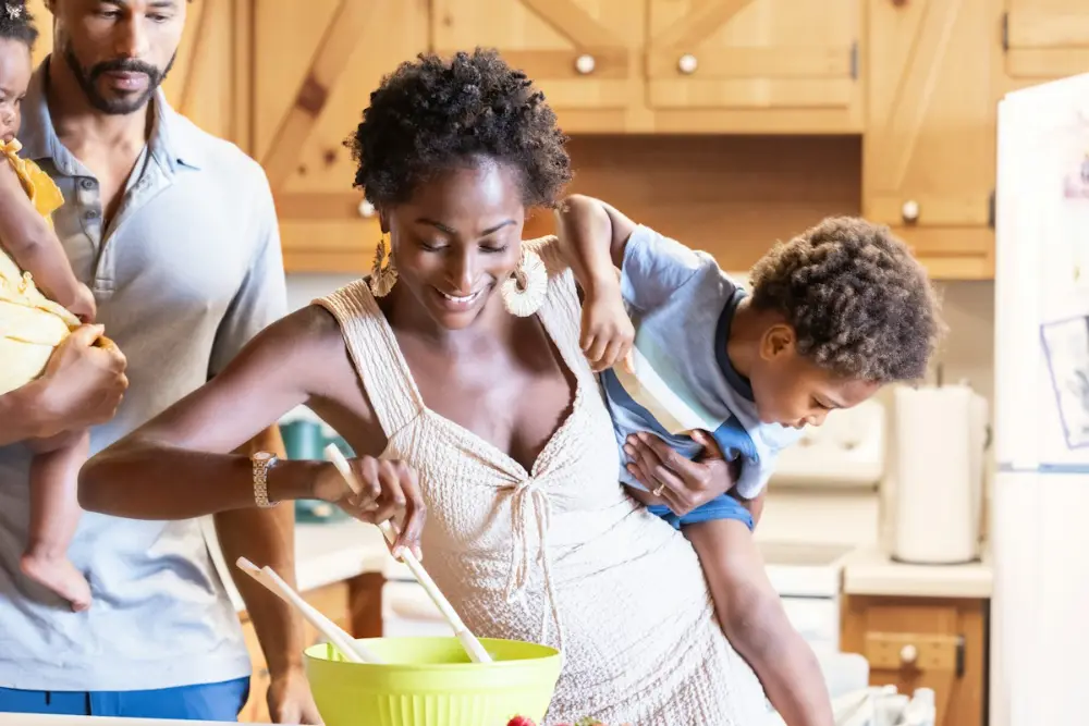 Una familia cocina en una cocina luminosa: la madre remueve en un bol verde con un niño a la espalda y el padre lleva a un bebé.