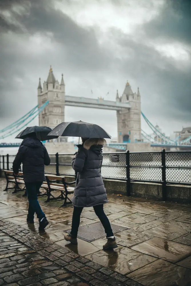 Dos personas con abrigo caminan bajo paraguas negros junto al río en un día de lluvia; al fondo se ve el Tower Bridge de Londres.