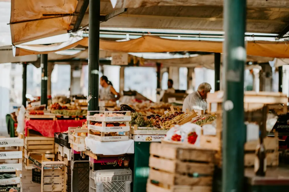 Un mercado cubierto con toldos naranjas y cajas de madera llenas de fruta; al fondo, un vendedor mayor y una mujer.