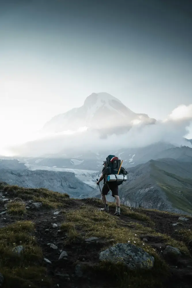 Un excursionista con una mochila grande sube por una ladera de montaña; al fondo, una montaña nevada entre nubes.