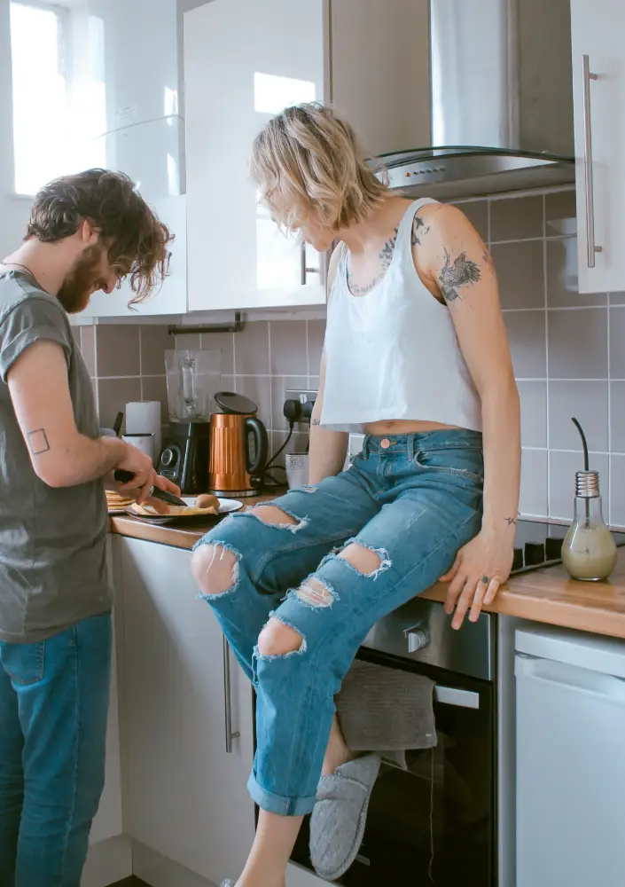Un chico con barba prepara comida junto a la encimera mientras una chica con vaqueros rotos está sentada encima de ella.