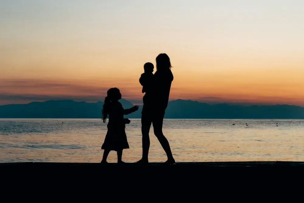 Siluetas de una mujer con un niño en brazos y una niña junto al mar al atardecer, con el cielo naranja y montañas al fondo.