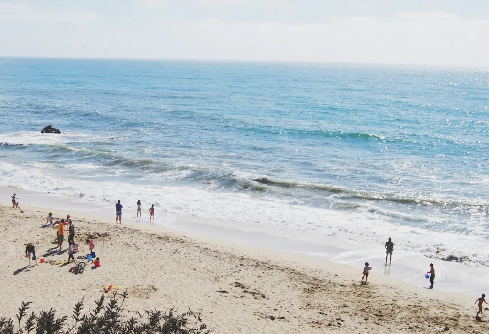Vista desde lo alto de una playa de arena con el mar azul y olas; varias personas y niños juegan en la orilla.