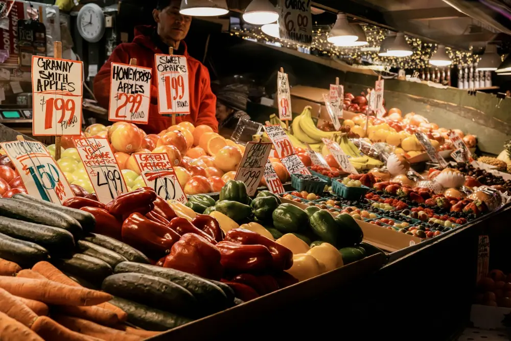 Un puesto de mercado lleno de frutas y verduras con carteles de precios; detrás, un vendedor con sudadera roja.