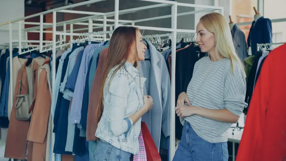 Dos mujeres jóvenes sonríen y hablan entre percheros de ropa en una tienda; una lleva una bolsa roja.