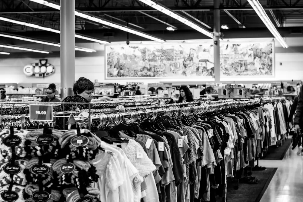 Foto en blanco y negro de una tienda grande de ropa de segunda mano con percheros llenos y algunas clientas mirando.