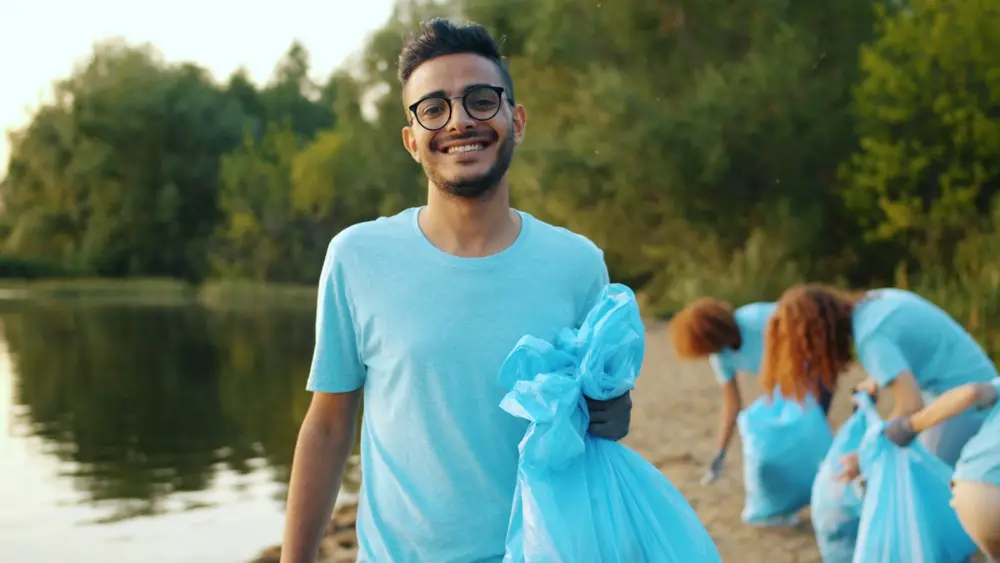 Un chico joven con gafas sonríe a la cámara con una bolsa azul de basura; detrás, dos mujeres recogen basura junto a un lago.